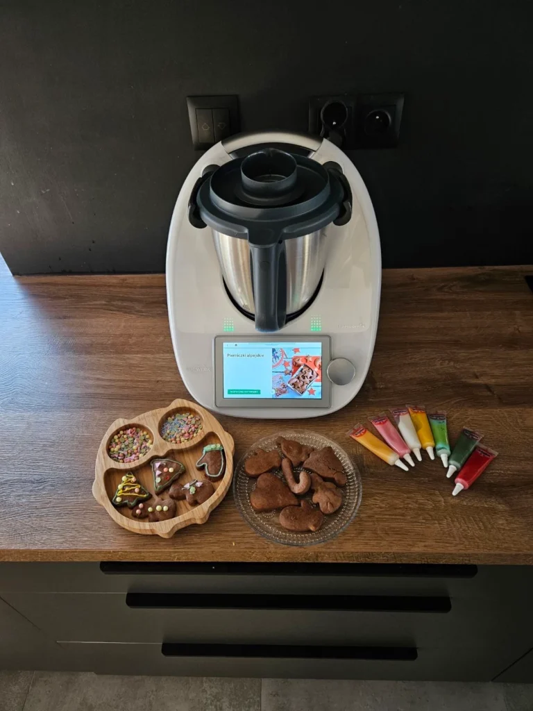 Thermomix TM6 na drewnianym blacie w kuchni, otoczony dekorowanymi piernikami, kolorowymi pisakami cukrowymi i posypkami, gotowy do świątecznych wypieków.