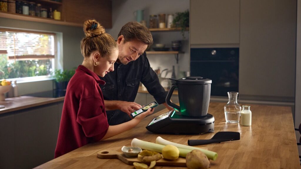 Mężczyzna i dziewczynka korzystają z Thermomixa TM7 w kuchni, obsługując urządzenie za pomocą smartfona. Nowoczesne gotowanie z Thermomixem 2025.